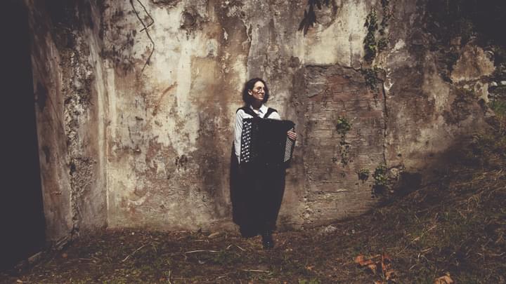 A woman standing in front of an old distressed plaster wall holding an accordion