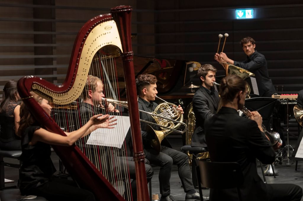 A mixed musical ensemble in rehearsal, viewed through the strings of a harp