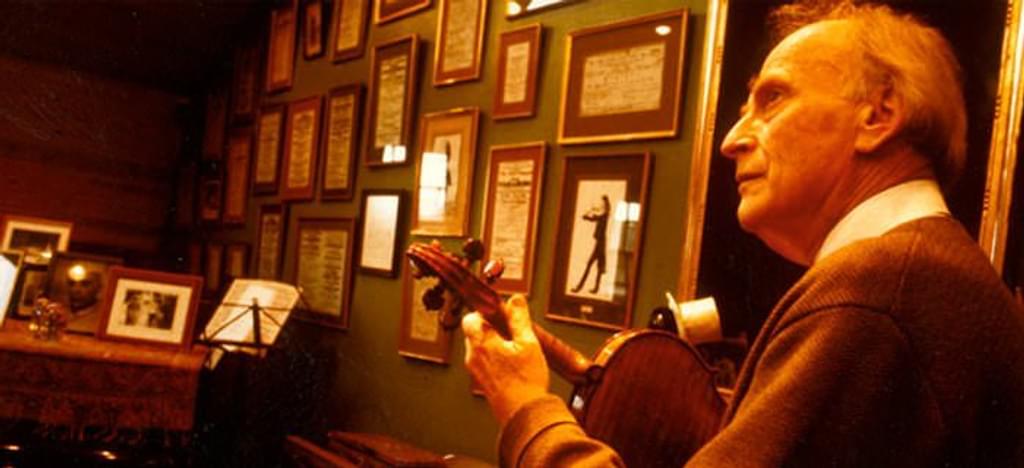 A violin player viewed from behind their left shoulder, plucking the instrument in front of a wall of framed certificates