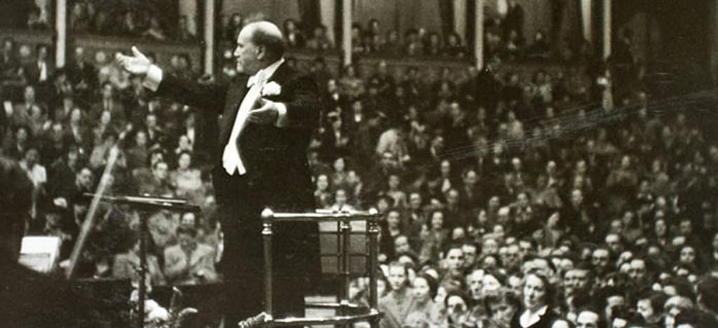 Scanned black and white photo of Sir Henry Wood stood atop a conductors podium, with raised arms in front of a busy concert audience.