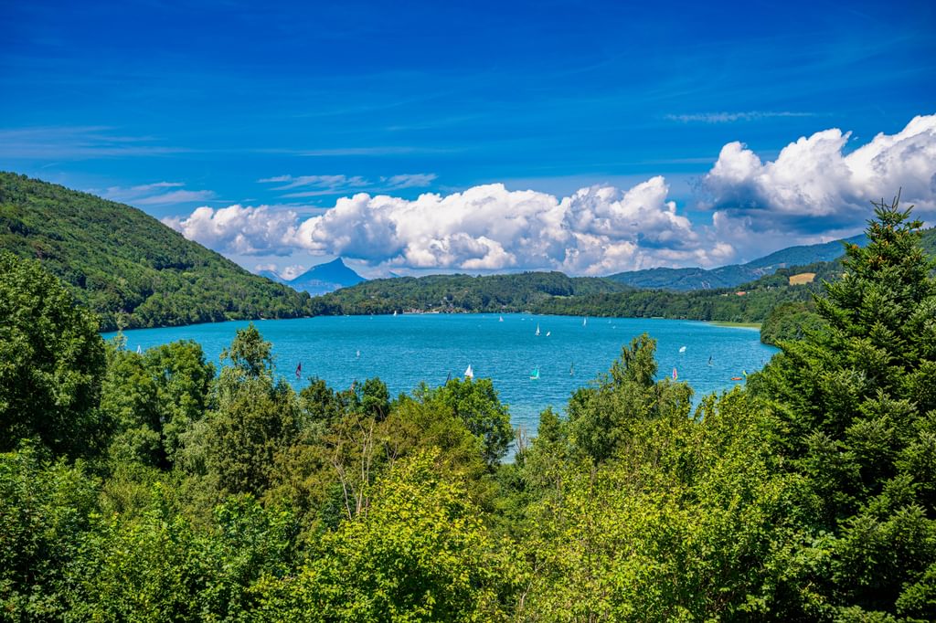 Landscape image of a blue lake surrounded by trees and mountains with blue sky and white clouds