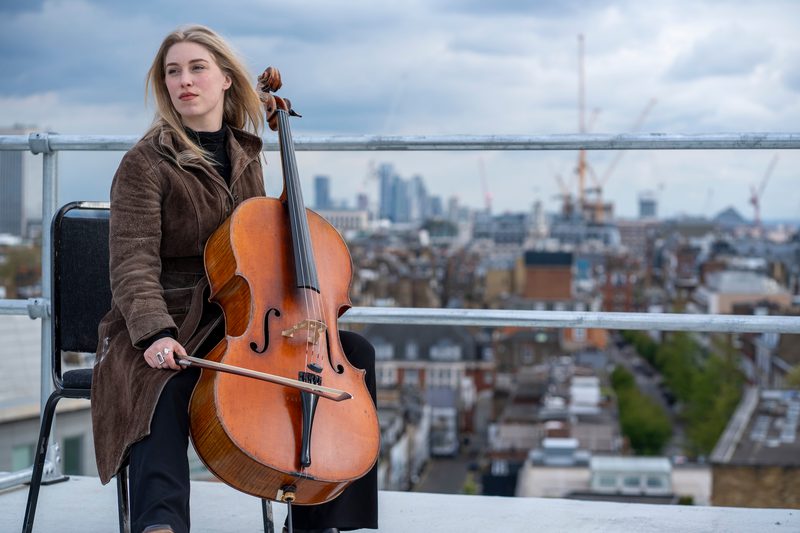 A cello student sits casually on a rooftop with a cello