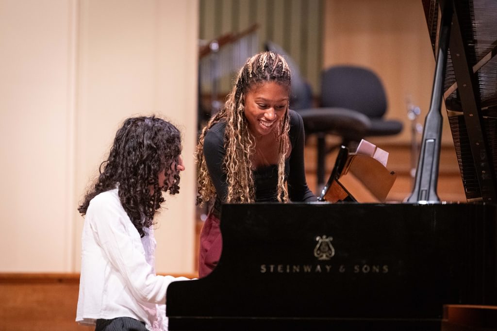 Isata Kanneh-Mason standing over a piano, smiling, with a student sitting at the piano
