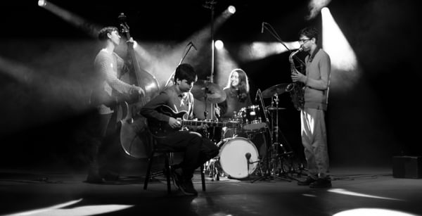 A band of four musicians performs in black and white, lit by spotlights and a smokey background