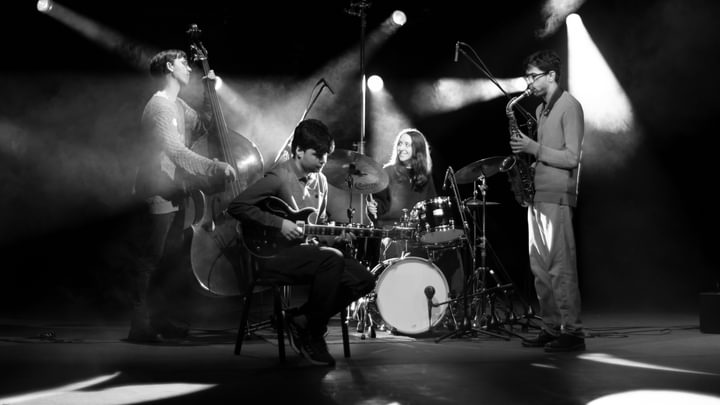 A band of four musicians performs in black and white, lit by spotlights and a smokey background