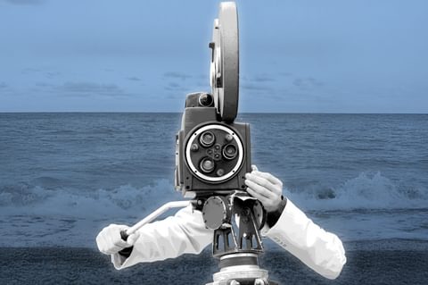 In black and white, the disembodied arms of a filmmaker in a lab coat operate an old-style film camera, with a backdrop of the grey sea
