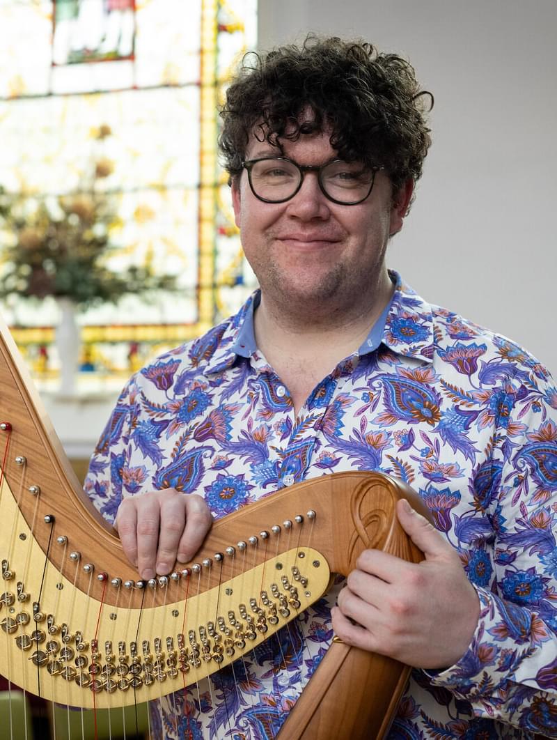 Alexander Rider smiels for camera, holding a harp