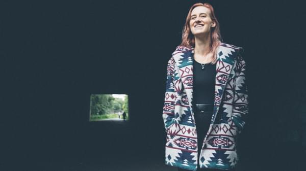 A woman standing smiling in a brightly coloured patterned coat against a dark background, with a small square of light behind from a tunnel opening