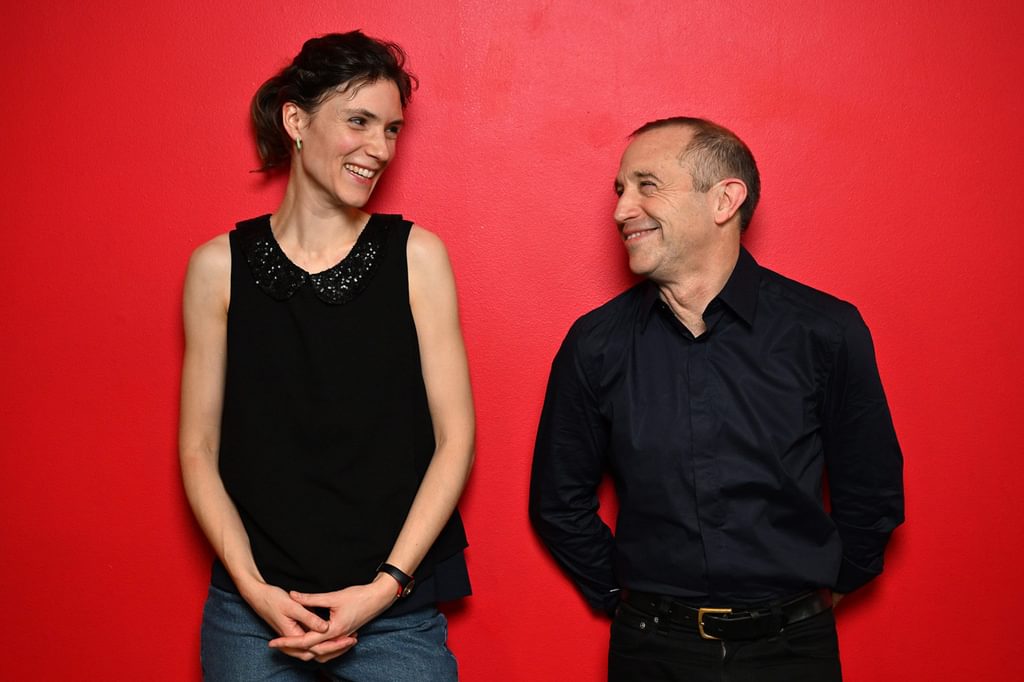Two people lean against a red wall, smiling to each other