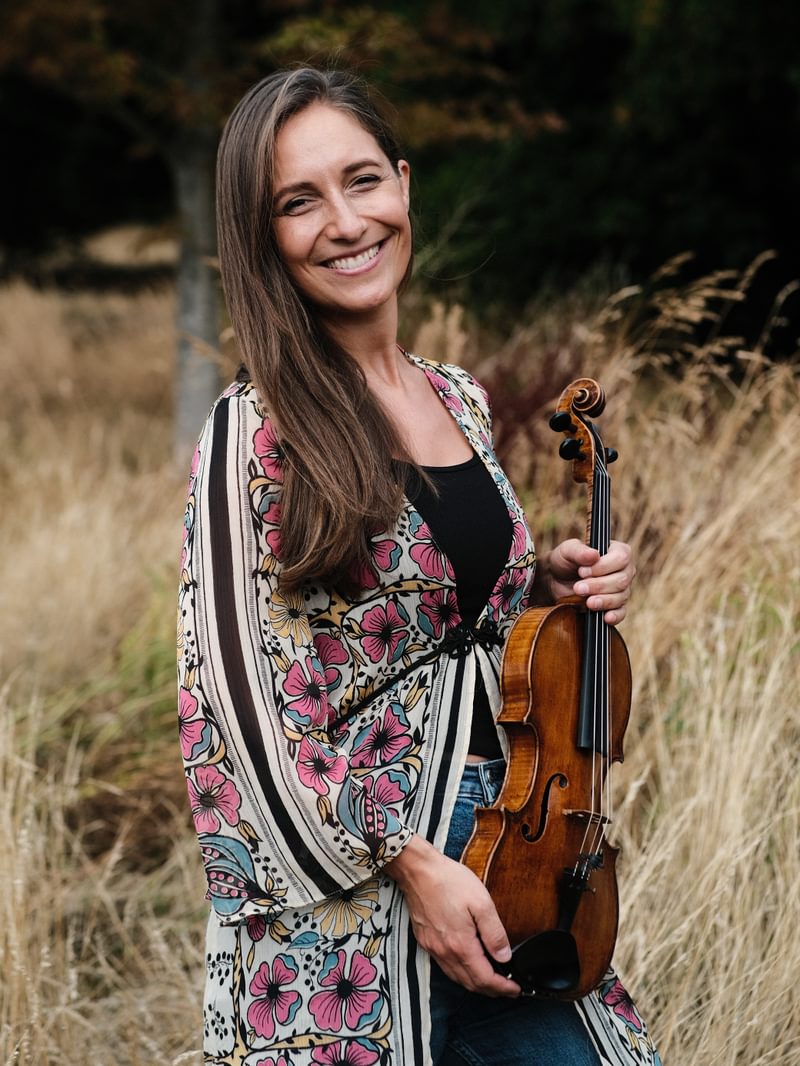 Portrait of Charis Jenson in a rural outdoor setting, holding a violin at waist height.