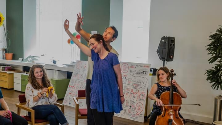 A group of seated and standing workshop participants smile and make expressive hand gestures, accompanied by a cellist.