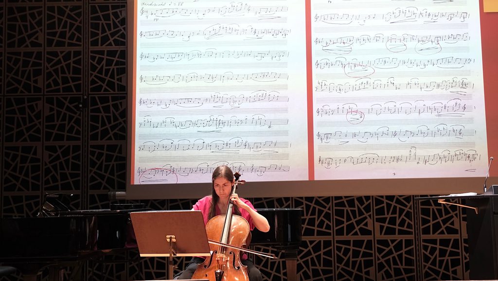 A cellist performs on stage in front of a large projection of sheet music