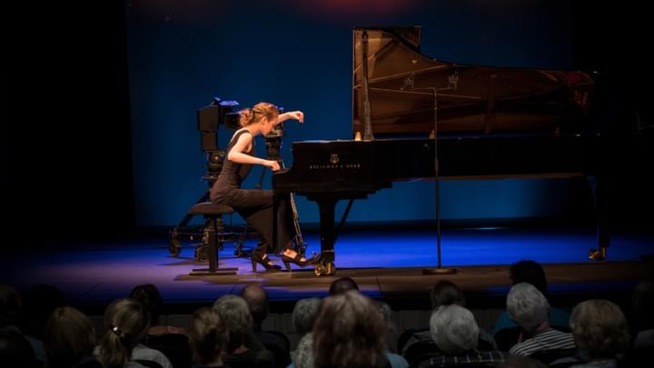 A woman on stage playing a grand piano for a concert audience, under a blue spotlight