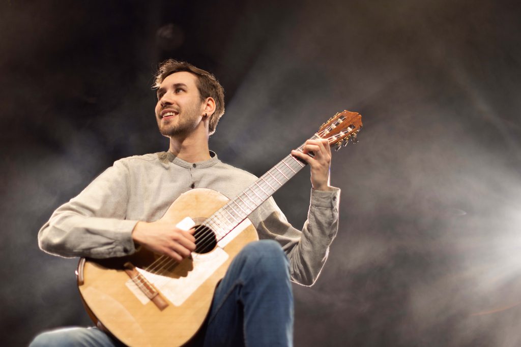 Gonçalo Maia Caetano playing a classical guitar and looking off into the distance whilst smiling. In the background there are stage lights and fog.