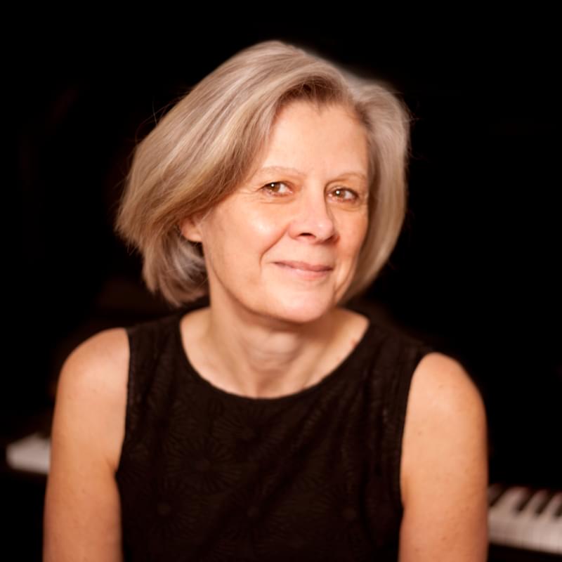 Helen Cawthorne sits at a piano, smiling for camera against a black background