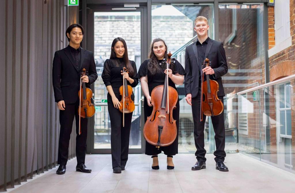 A string quartet smile to camera, holding their instruments outside a concert hall