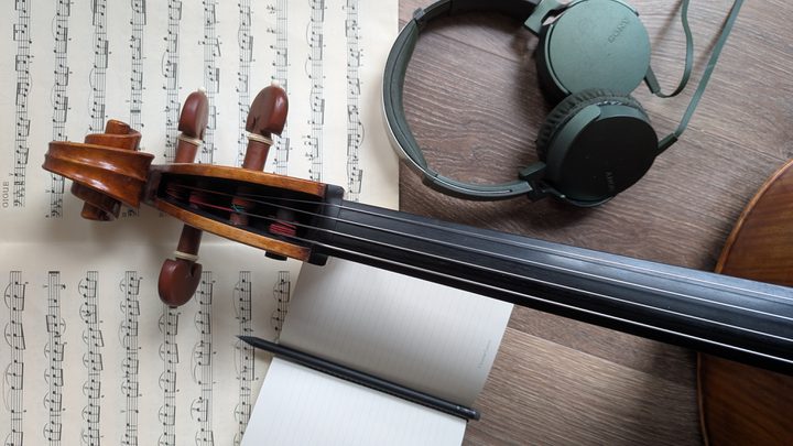 A close-up of the neck of a violin, with a manuscript, a notepad and some headphones