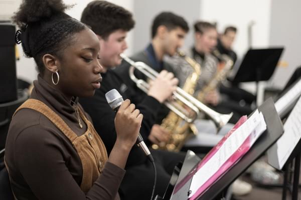 A young vocalist sings into microphone while a brass section performs in the background