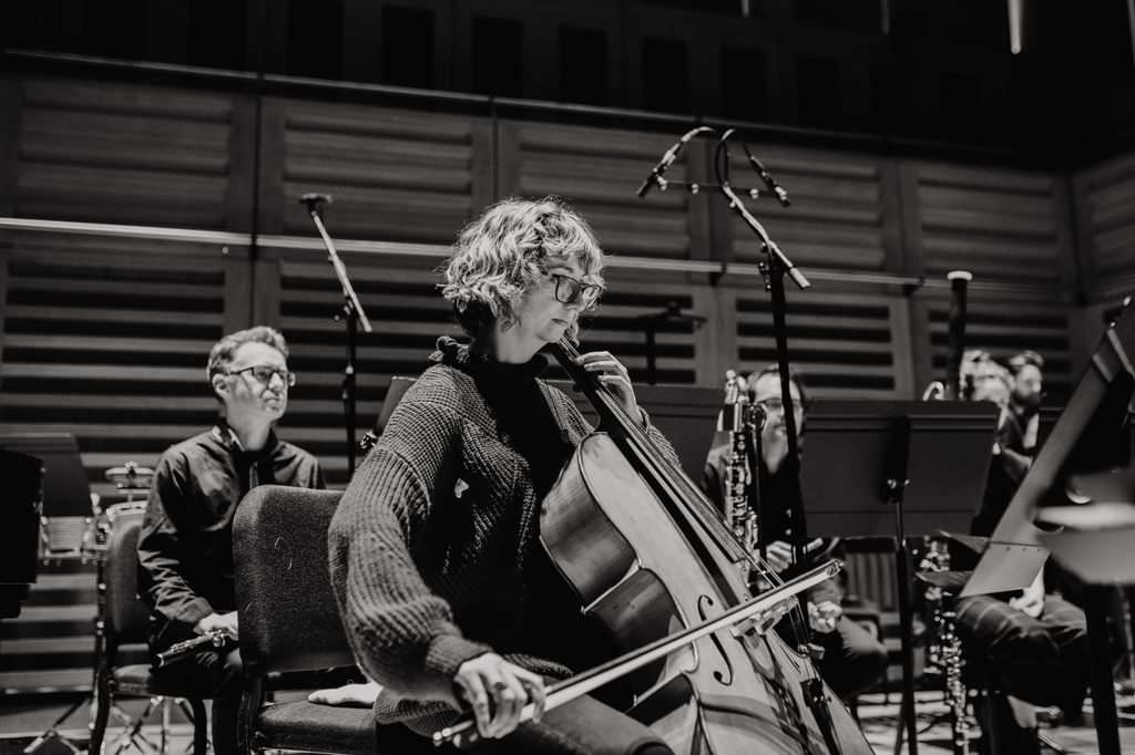 Black and white image of a woman seated with a cello, on stage as part of an ensemble