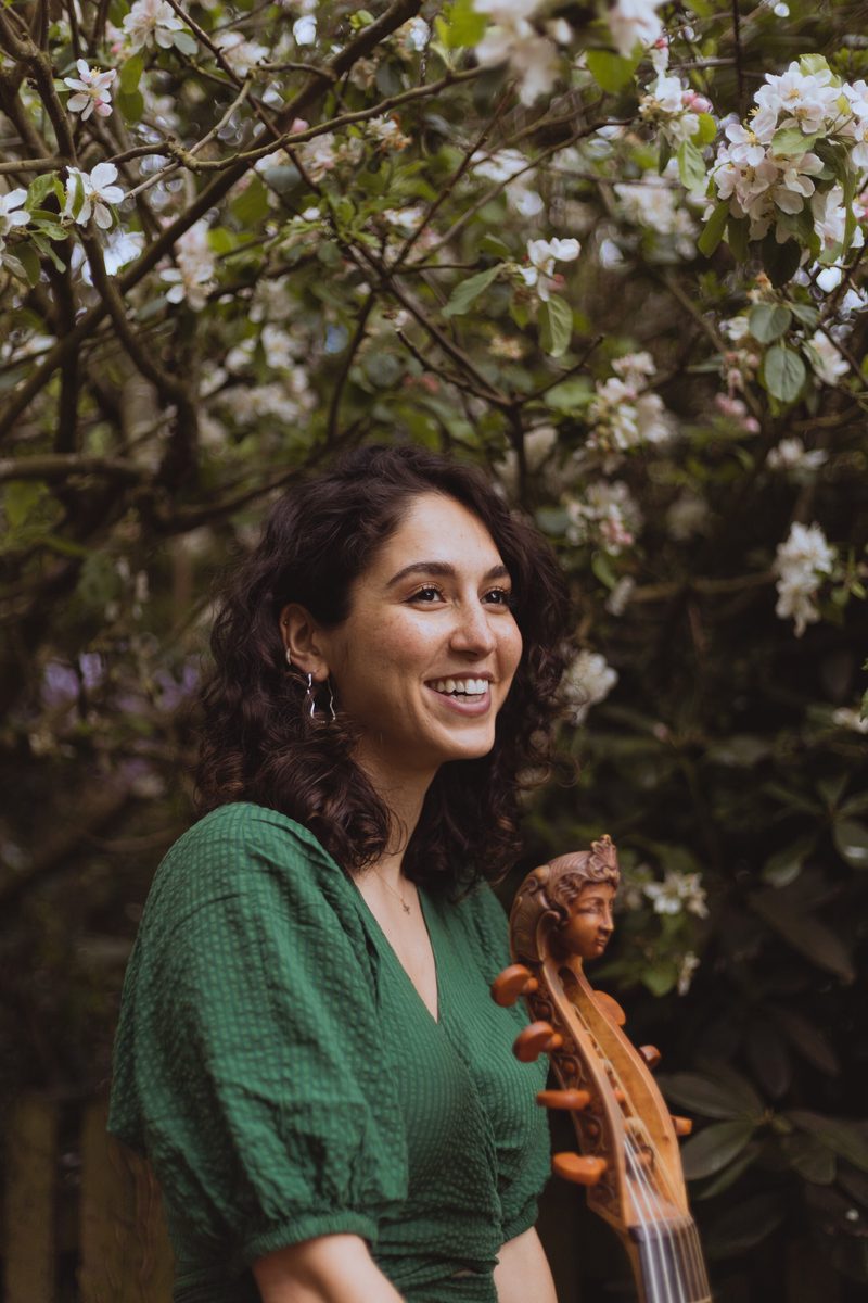 A woman in a green dress, pictured smiling, with the neck of a stringed instrument in the foreground and a tree in the background