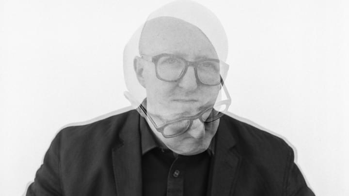 Black and white headshot of a man facing the camera wearing dark framed glasses with a superimposed headshot looking downwards