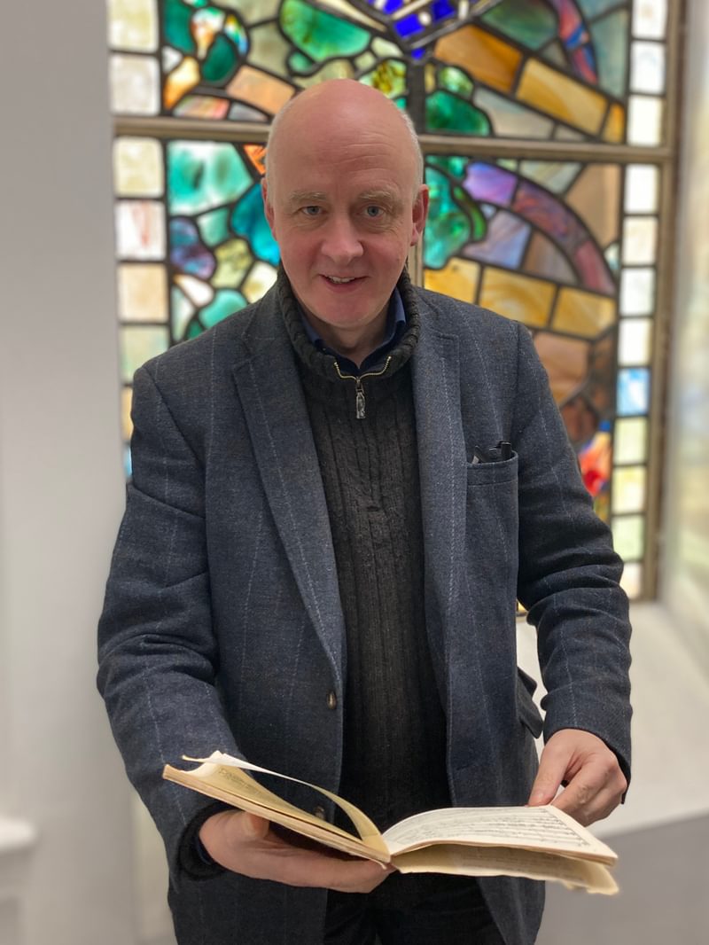 Matthew Taylor poses for camera in front of a stained glass window, holding a book