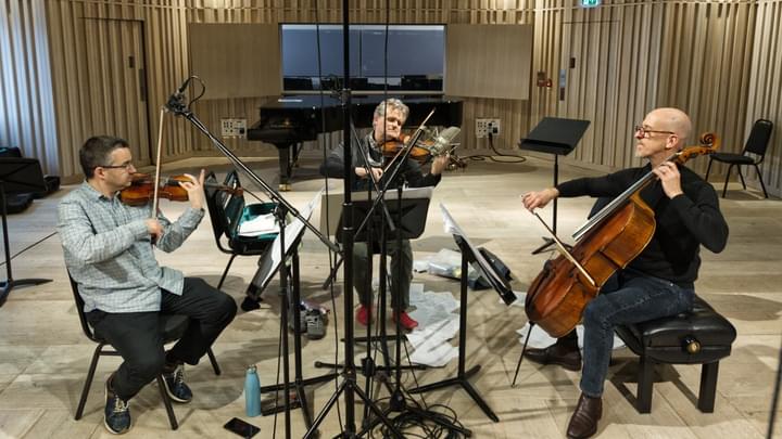 Three musicians sitting together in a semi-circle in a studio playing violin, viola and cello, with microphones and cables to record their sound