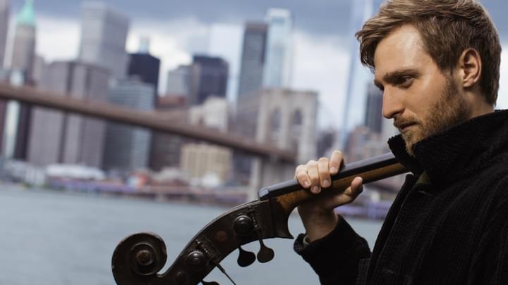 A side profile of a man holding a double bass over his shoulder, the scroll and pegs in front of him, with a city river background.