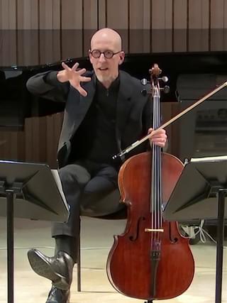 A man seated with a cello and bow supported in one hand, gesturing to speak with the other hand.
