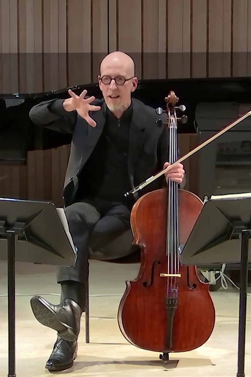 A man seated with a cello and bow supported in one hand, gesturing to speak with the other hand.