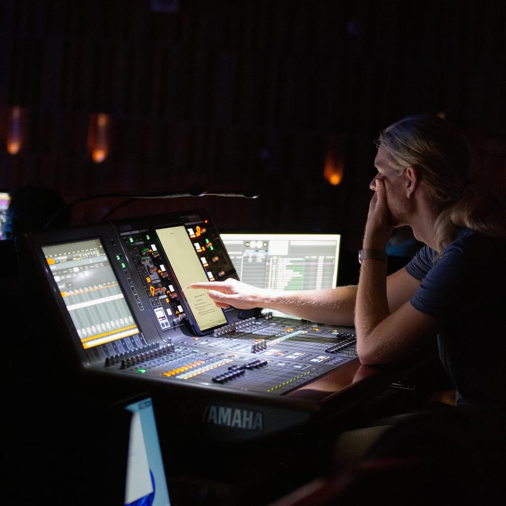 A sound engineer sits at a technical desk working on a touch screen device in a dark auditorium
