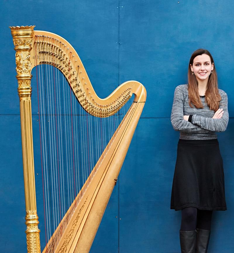 Rachel Wick stands against a blue background next to a harp, crossing arms