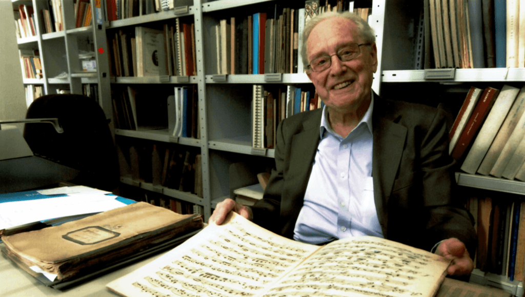 Portrait of Sir Charles Mackerras sitting at a desk, smiling to camera while leafing over a manuscript.