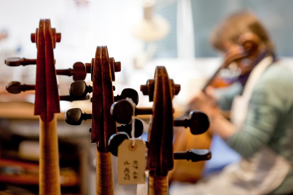 A person plays a violin out of focus in the background, with a row of violins on a rack in the foreground