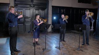 Four standing brass players perform in a wooden-panelled room.
