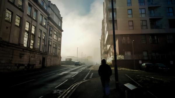 A person in shadow walking between two tall buildings separated by a city road with a misty cloud between them in the distance.