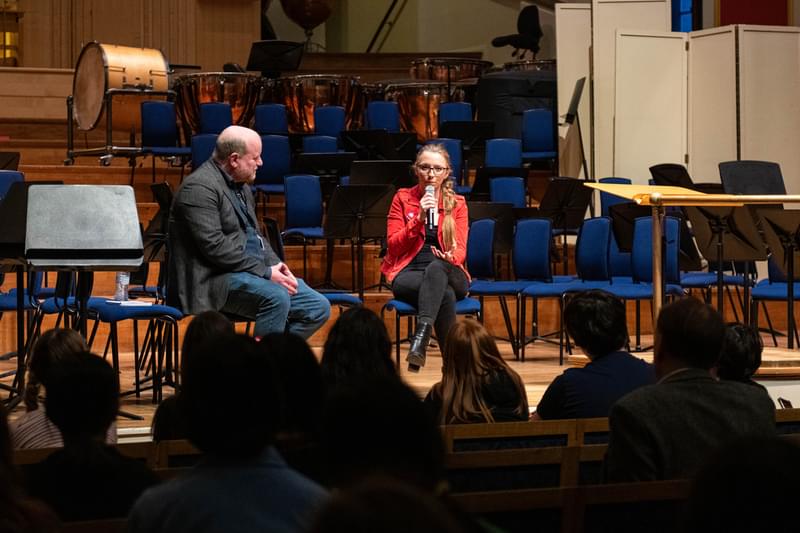 Anna Lapwood on the stage of the Duke's Hall, holding a microphone and addressing a room of Junior Academy students