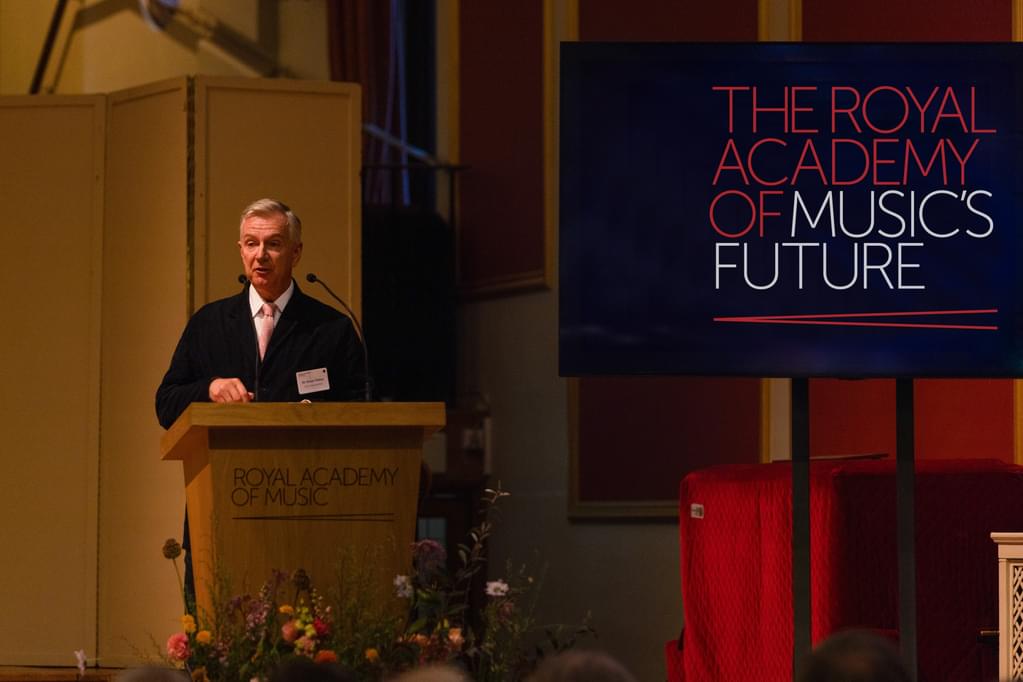 Sir Simon Robey talks at a lecturn, alongside a screen with the text 'The Royal Academy of Music's Future'