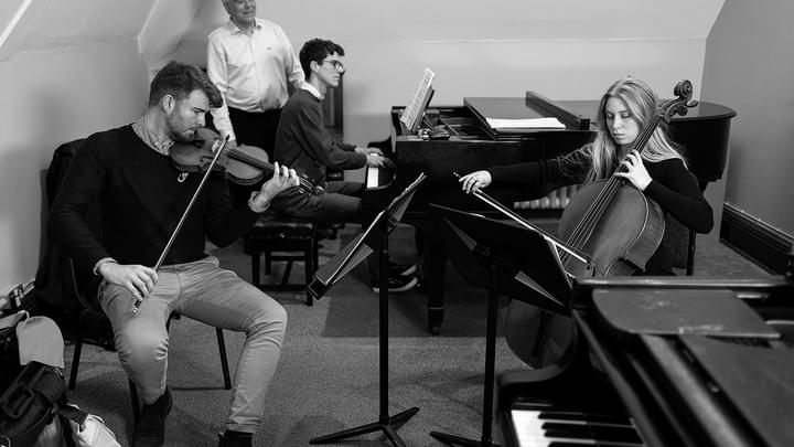 An ensemble piano tutor guides a piano accompanist in a practice room with a string duet (black and white)