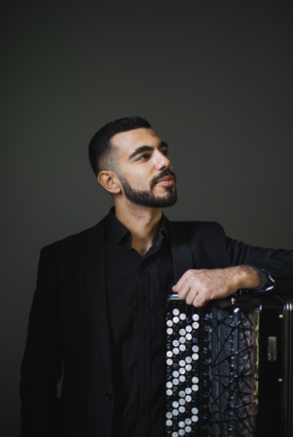 Portrait image of Flavio Rodrigues looking off to the right, leaning his hand on an accordion