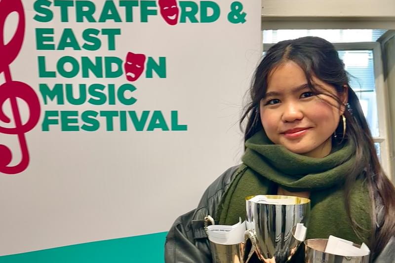 An image of Junior Academy Musical Theatre student Kaye Lee holding three trophies, standing in front of a banner for the Stratford and East London Music Festival