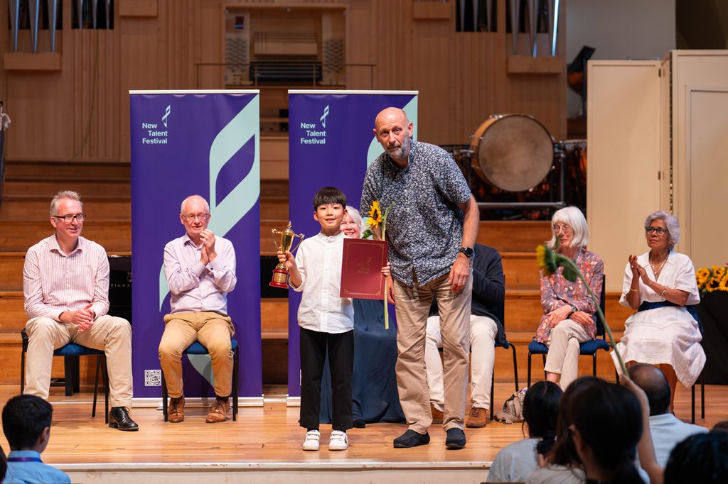 A young musician being presented an award on a stage, with several adults sat behind them applauding