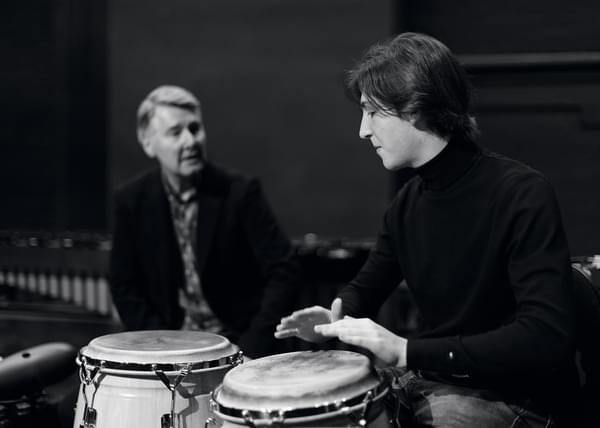 A student hand playing a conga drum with a tutor in the background (black and white)
