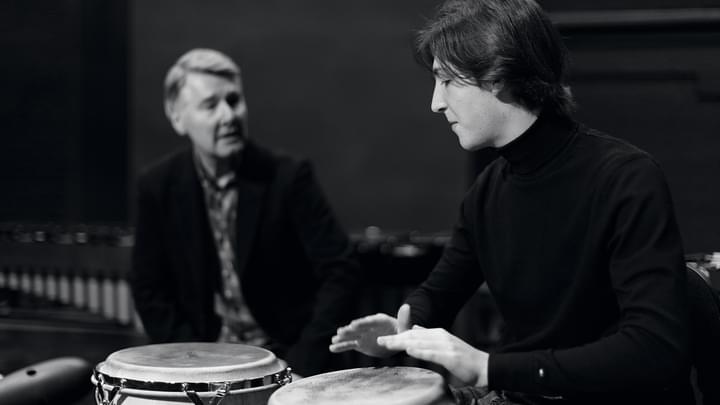 A student hand playing a conga drum with a tutor in the background (black and white)