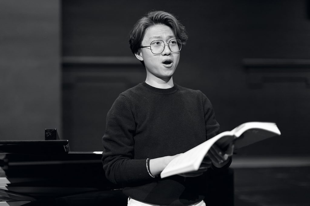 A vocal studies student holds sheet music in hand while looking out of frame, singing (black and white)
