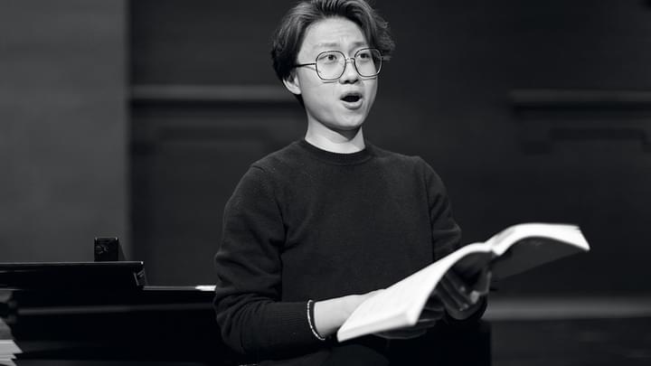 A vocal studies student holds sheet music in hand while looking out of frame, singing (black and white)