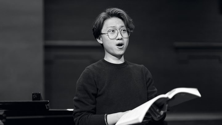 A vocal studies student holds sheet music in hand while looking out of frame, singing (black and white)