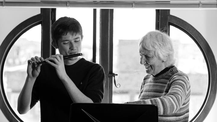 A student plays a piccolo while being guided by a tutor to sheet music (black and white)