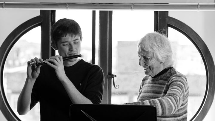 A student plays a piccolo while being guided by a tutor to sheet music (black and white)
