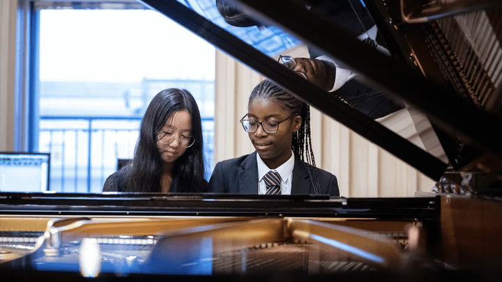 Two young students sit at a piano keyboard, smiling